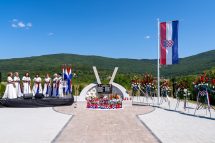Memorial plaza with Croatian flag, flower wreaths, and a central curved monument; choir in white dresses on the left.