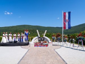 Memorial plaza with Croatian flag, flower wreaths, and a central curved monument; choir in white dresses on the left.