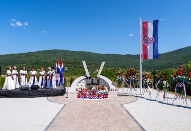 Memorial plaza with Croatian flag, flower wreaths, and a central curved monument; choir in white dresses on the left.
