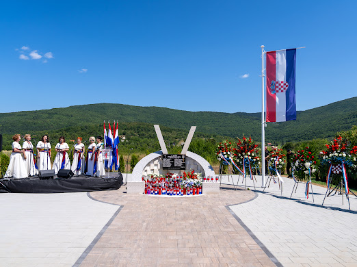 Memorial plaza with Croatian flag, flower wreaths, and a central curved monument; choir in white dresses on the left.