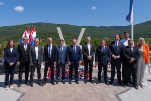 Group of formally dressed officials posing for a ceremony photo with flags and a memorial sculpture, mountains in the background.