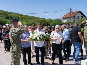 Casketless memorial: a man in white shirt carries a large white floral arrangement as others watch; soldiers salute in a ceremony outdoors.
