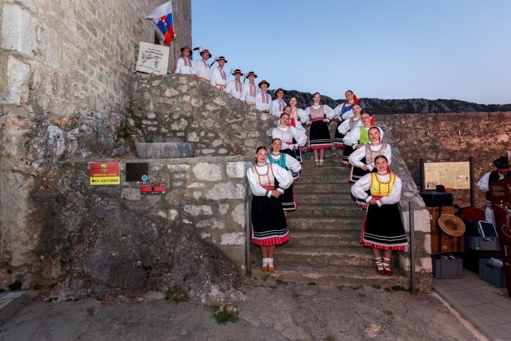 Group of people in traditional Eastern European folk costumes posing on stone fortress steps at dusk.