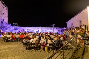 Outdoor nighttime concert in a stone-walled courtyard, with a large audience seated facing a small stage.
