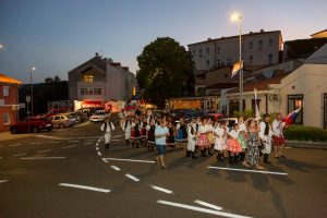 People in traditional folk costumes marching in a street parade at dusk, with flags and onlookers along the roadside stores and cars parked nearby.