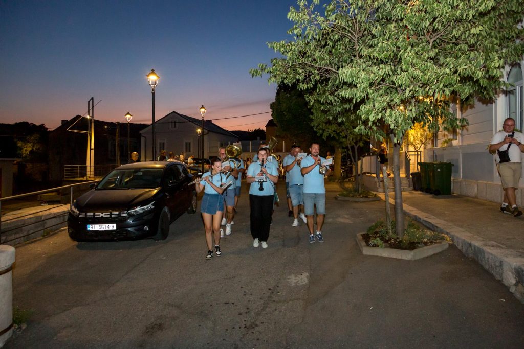 A small street parade at dusk with a blue-shirt band playing wind instruments and drums, walking toward the camera.