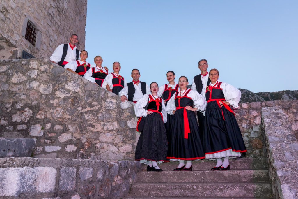 Group of people in black and red traditional folk costumes posing on stone steps beside a castle wall.