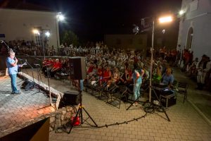 Man speaks into a microphone on a small stage at night, audience seated in courtyard with buildings and bright lights behind.