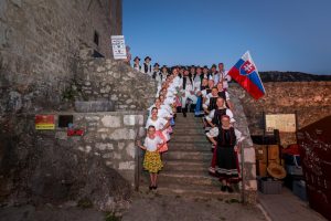 Group of people in traditional Slovak folk costumes posing on stone castle stairs, with a Slovak flag in the background.