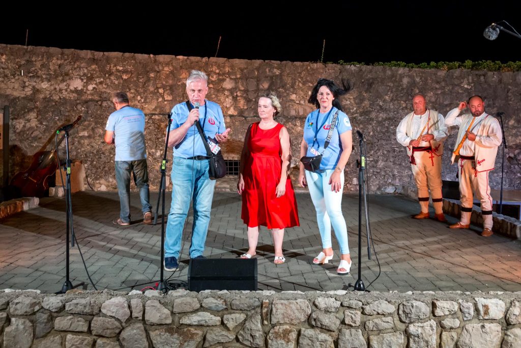 Group of six performers on an outdoor night stage, microphones set up, stone wall behind them.