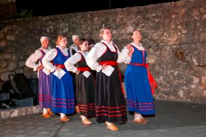 Group of women in traditional folk costumes performing a dance in front of a stone wall at nightfall.