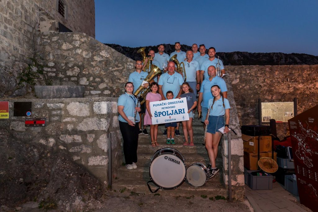 Group of musicians in light blue polo shirts posing on stone steps with brass instruments outdoors at dusk; a sign held by children reads 'PUHAČKI ORKESTAR PETROVSKO ŠPOLJARI CROATIA'.