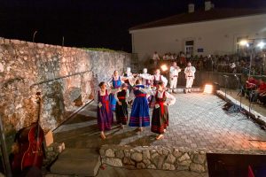 Nighttime outdoor folk dance performance with dancers in traditional blue and red costumes and musicians on a stone courtyard stage, audience watching behind a wall.