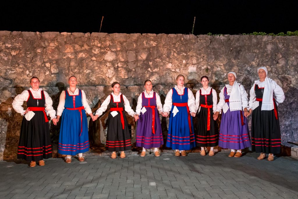 Nine women in colorful traditional dresses stand hand in hand against a stone wall at night.