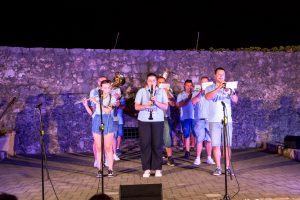 Nighttime outdoor brass band performing in front of a stone wall, musicians reading sheet music.
