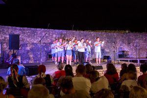 Group of musicians in light blue shirts perform on an outdoor stage with a stone wall backdrop at night; audience watches.