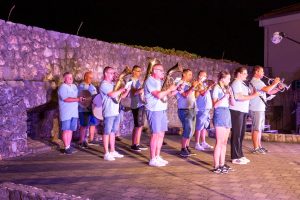 Nighttime outdoor band performance with musicians in light-blue shirts against a stone wall; brass and wind instruments in use.",