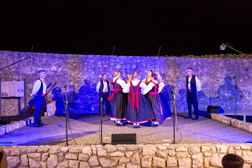Group of dancers in traditional folk dresses form a circle on an outdoor stage at night, with a stone wall and blue lighting in the background.