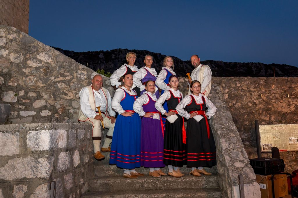 Group of performers in colorful traditional costumes posing on ancient stone steps at night, with a ruined wall behind.