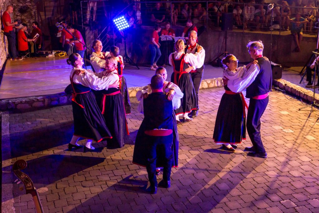 Pairs of dancers in traditional red-and-black outfits dance on a cobblestone courtyard at night, with a lit stage and audience in the background.
