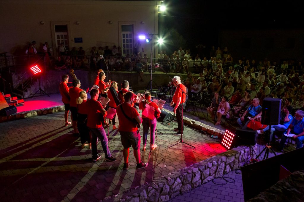 Nighttime outdoor concert with a red-clad choir performing on a stone stage, audience seated behind a low wall.