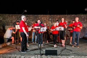 Group of musicians in red shirts performing with guitars on an outdoor night stage in front of a stone wall.