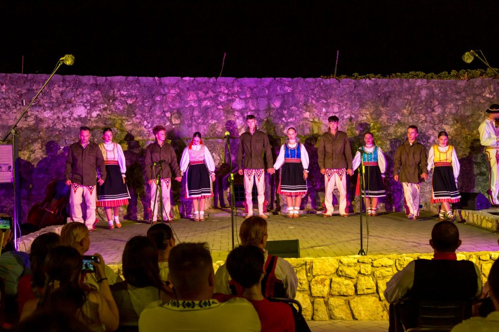 Group of young performers in traditional folk costumes standing in a line on an outdoor stage at night, holding hands, with a purple-lit stone wall behind them and an audience in the foreground.