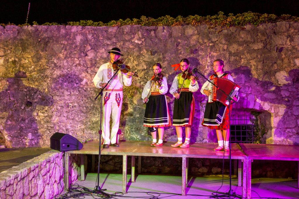 Five musicians in traditional folk costumes perform on a stone outdoor stage at night; a man plays violin on the left while four women, two with violins and one with an accordion, stand and play under purple lighting.