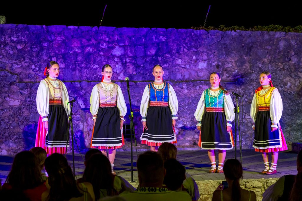 Five women in traditional folk costumes perform on a night stage in front of a stone wall, microphones nearby.