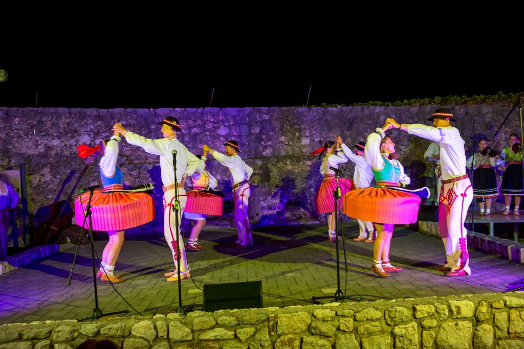 Dancers in white shirts and colorful striped skirts perform a circle dance on a stone stage at night, with musicians in the background against a stone wall.
