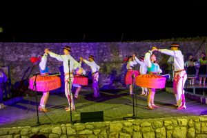 Dancers in white shirts and colorful striped skirts perform a circle dance on a stone stage at night, with musicians in the background against a stone wall.