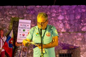 Man in a teal shirt speaks into a microphone at a nighttime outdoor event, holding notes with a lanyard badge visible.