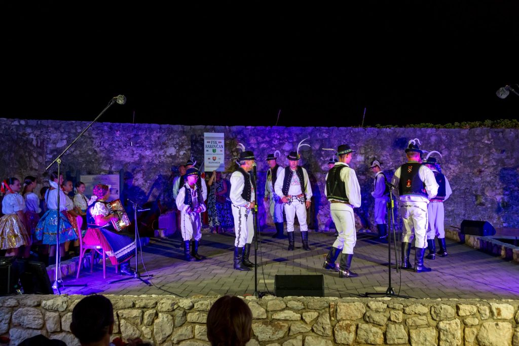 Nighttime folk performance on a stone-walled stage; dancers in traditional clothing and musicians with instruments under blue-purple lighting.