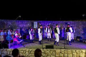 Nighttime folk performance on a stone-walled stage; dancers in traditional clothing and musicians with instruments under blue-purple lighting.