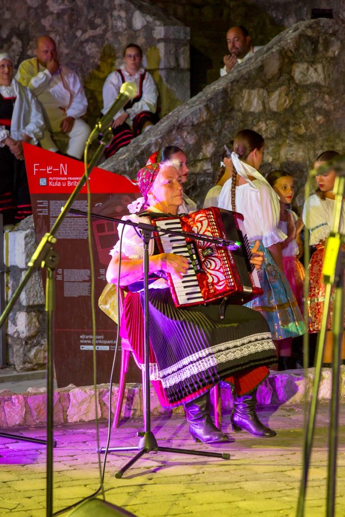An elderly woman in a colorful traditional outfit plays a red accordion on a stone stage with microphones around her.