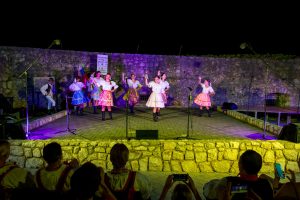 Group of dancers in white blouses and colorful skirts performing a folk dance on an outdoor stage at night, with a stone wall backdrop and audience watching.