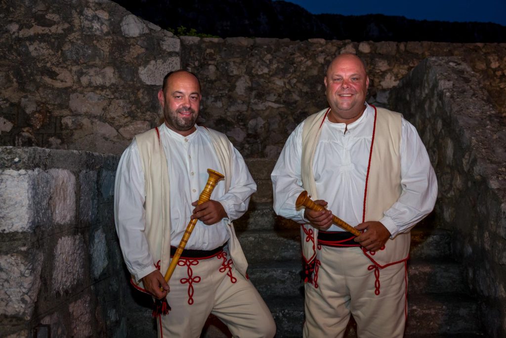 Two men in white traditional outfits with red embroidery, holding wooden wind instruments, standing in a stone ruin at night.