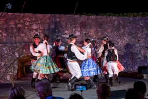 Group of people in traditional costumes dancing in a circle on a stone stage at night.