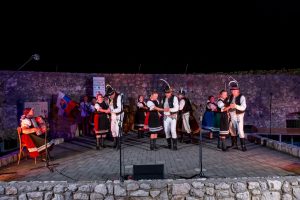 Folk dancers in traditional costumes perform on an outdoor stone stage at night, with a musician playing the accordion on the left.