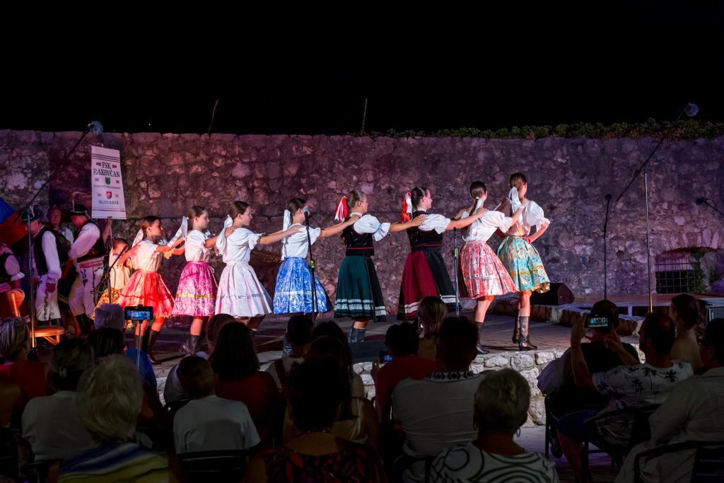 Group of girls in colorful traditional dresses performs a line dance on an outdoor stage at night beside a stone wall.
