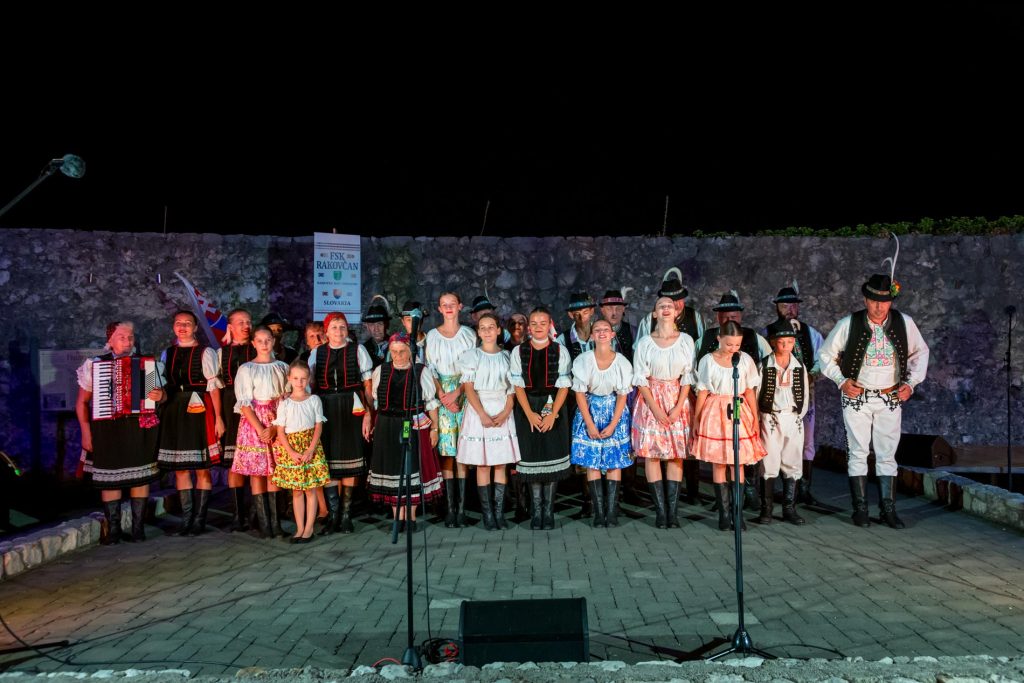Group of performers in colorful traditional folk costumes standing on a nighttime outdoor stage with microphones and a stone wall backdrop.