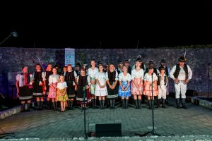 Group of performers in colorful traditional folk costumes standing on a nighttime outdoor stage with microphones and a stone wall backdrop.