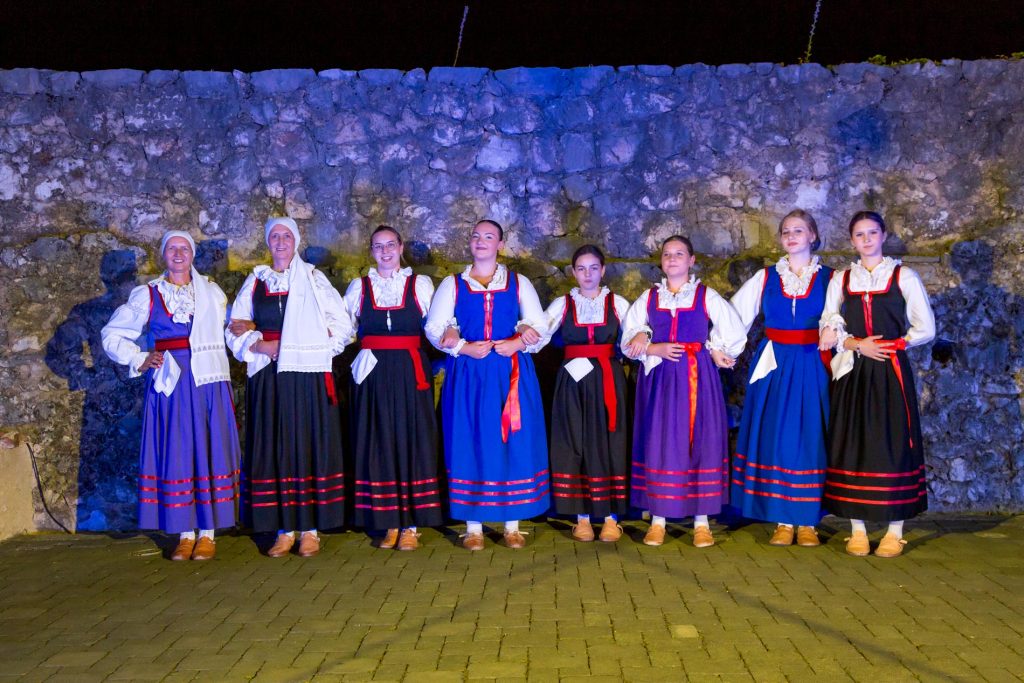 Nine women in traditional folk dresses with white blouses and red sashes stand arm-in-arm in front of a stone wall at night, illuminated by blue lighting.