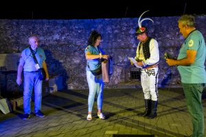 Man in traditional outfit with feathered hat presents a paper to a woman on a stage at night; others watch, blue lighting on stone wall behind.