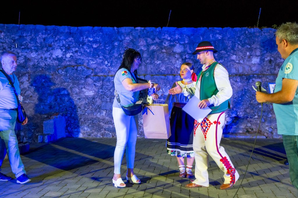 Woman in white sandals hands a gift bag to a man in a traditional green-vest folk costume on a blue-lit stage with a stone wall backdrop.