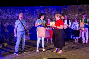 Two women exchange a gift on a night-time stage while others in traditional attire hold certificates and frames against a stone wall.