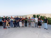 Group of people posing outdoors with flower wreaths and ribbons, mountains in the background (memorial/tribute scene).