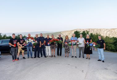 Group of people posing outdoors with flower wreaths and ribbons, mountains in the background (memorial/tribute scene).