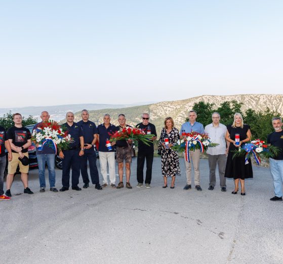 Group of people posing outdoors with flower wreaths and ribbons, mountains in the background (memorial/tribute scene).