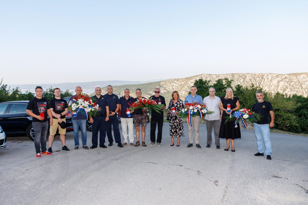 Group of people posing outdoors with flower wreaths and ribbons, mountains in the background (memorial/tribute scene).